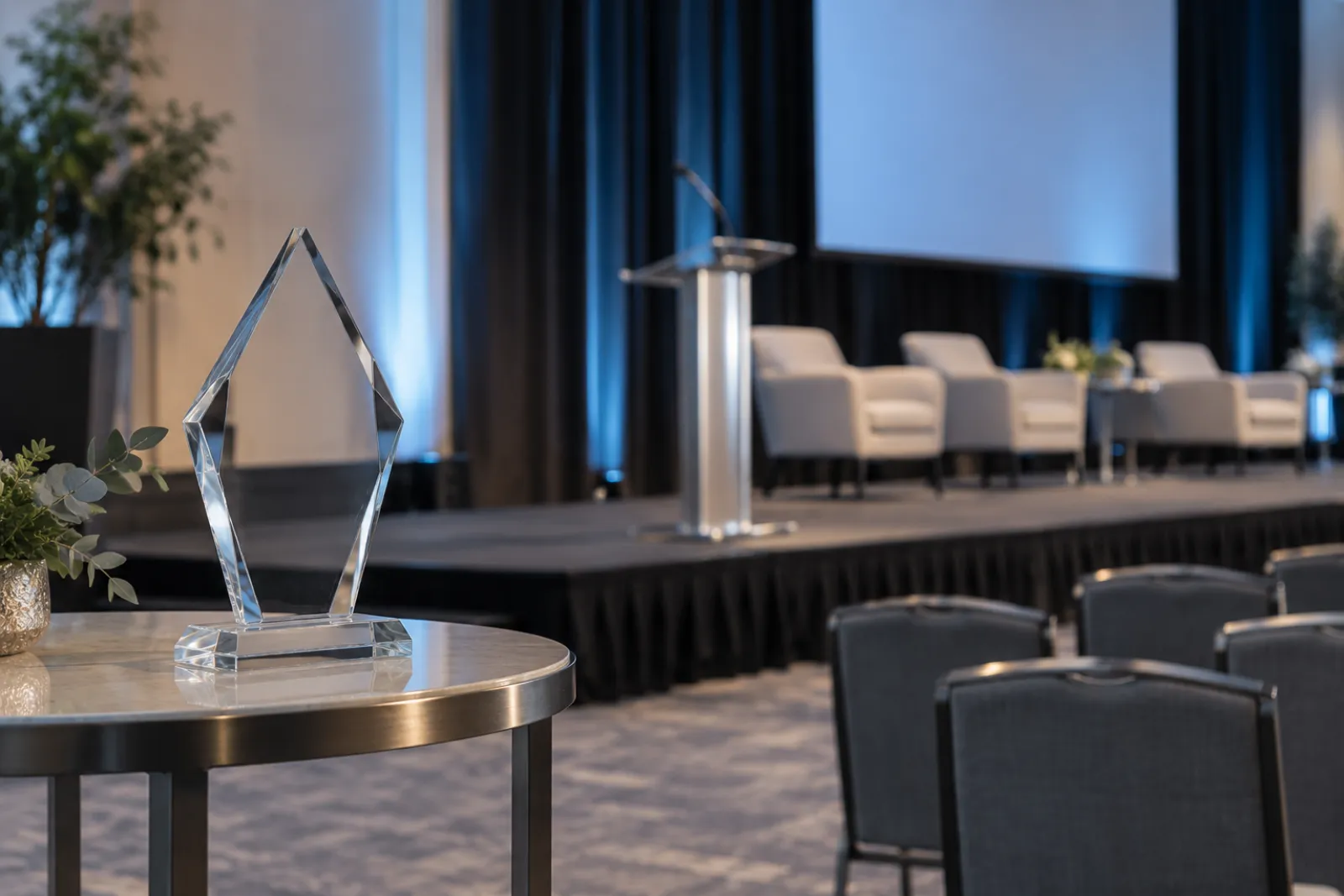 A generic clear glass award trophy with no engraving on a small side table near an empty conference stage, dim warm stage lighting, empty chairs blurred behind