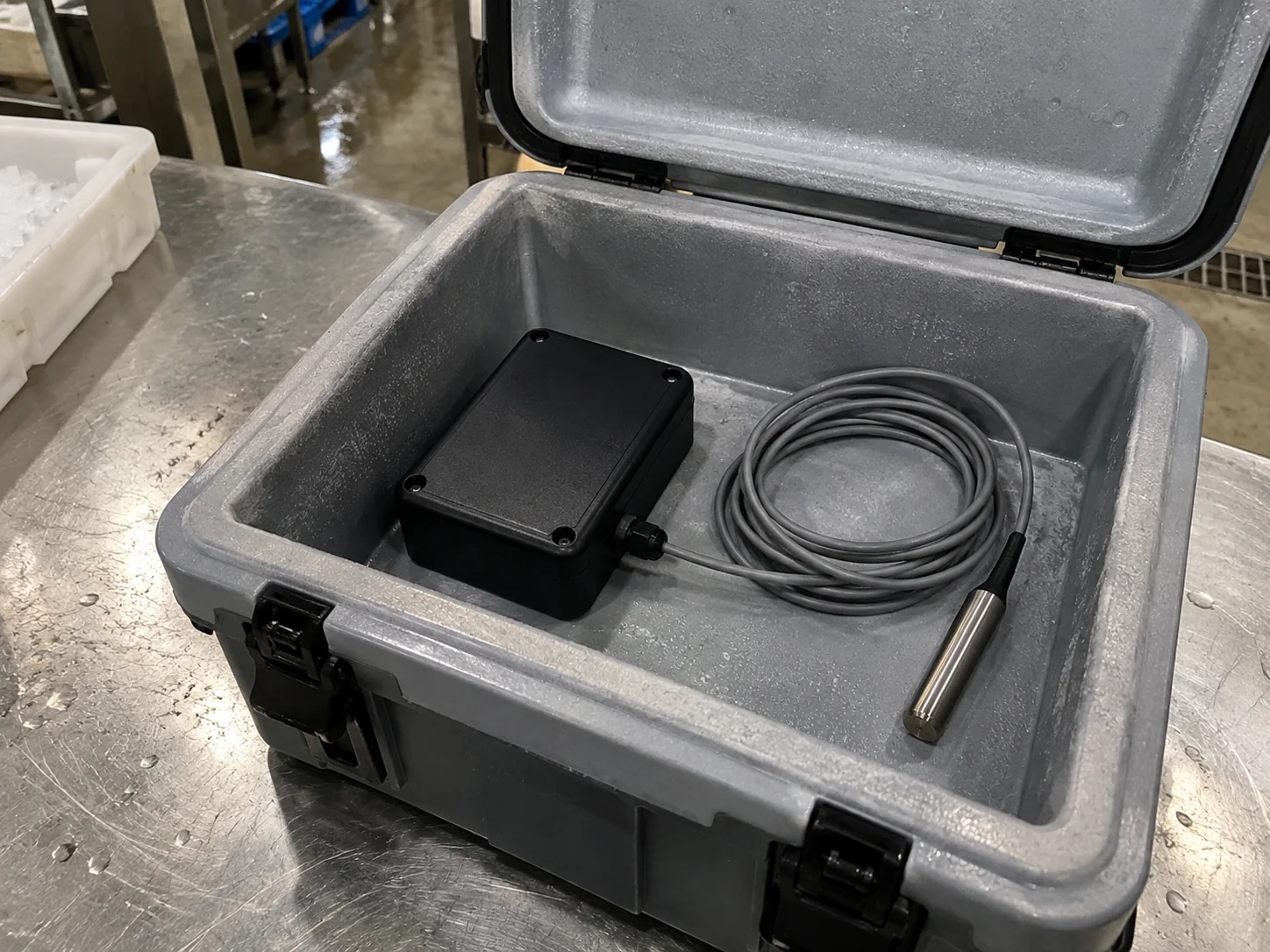 A matte-black temperature logger with an external stainless-steel probe resting inside an open insulated cold-chain container on a steel bench